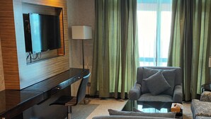 A hotel room with a flat-screen TV mounted on a wooden panel, a grey armchair, a patterned sofa, and a small table with a tissue box. - Hotel Al Shahba Jeddah   (Jeddah)