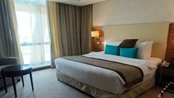 A hotel room with a large bed, a chair, a small table, and a window with curtains.