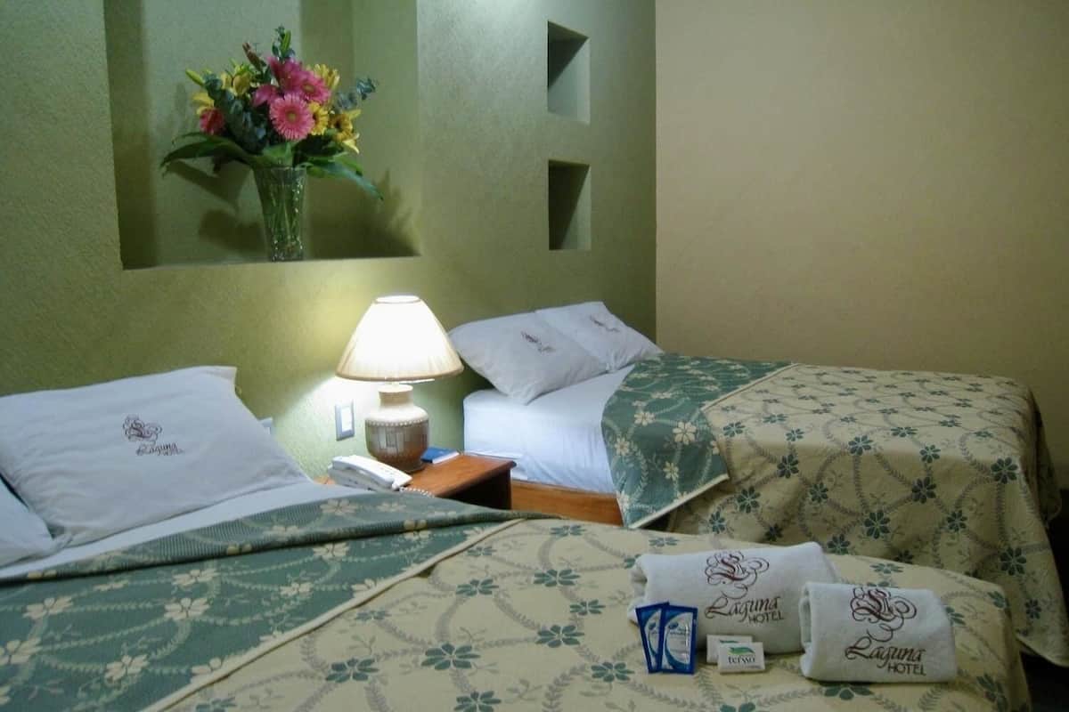 Habitación de hotel con dos camas, una mesita de noche con lámpara, un florero en la pared y una colcha con estampado floral.
