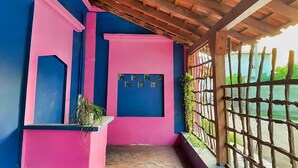 A room with a wooden ceiling, a pink and blue wall, a window with a view, and a small shelf with plants. - Hotel de Arnold (Mazatlán)