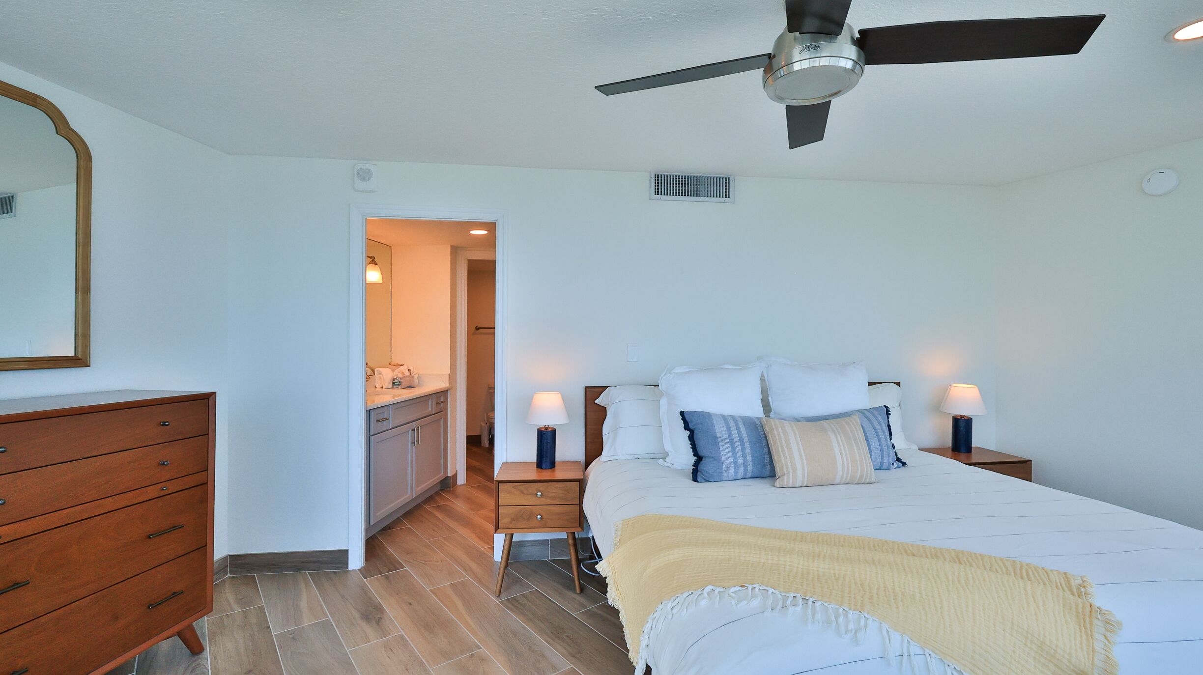 Un dormitorio con cama, mesitas de noche, ventilador de techo, cómoda y baño visible a través de una puerta abierta.