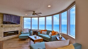 Una sala de estar con chimenea, ventanas amplias con vista al mar y un ventilador de techo.