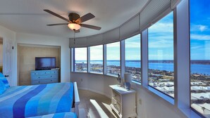 A bedroom with a large window, a ceiling fan, a television, and a bed with a blue quilt. - SEASHELL SERENADE-24th Floor Ocean/River View - POOL, HOT TUB, Peck Plaza 2B/2B (Daytona Beach)