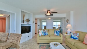 A living room with a beige sofa, a television, and decorative pillows. There is a ceiling fan and a view of the outside through a door. - SEASHELL SERENADE-24th Floor Ocean/River View - POOL, HOT TUB, Peck Plaza 2B/2B (Daytona Beach)