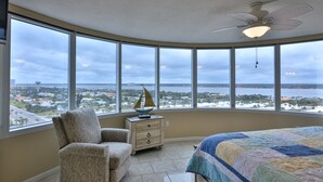 Un dormitorio con ventilador de techo curvo, un ventanal con vistas a un paisaje urbano y al agua, una silla, una mesita de noche y una cama con colcha.