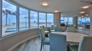 Amplia habitación con ventanas grandes que ofrecen vista a la playa y palmeras. Hay una mesa redonda con sillas y un ventilador de techo.