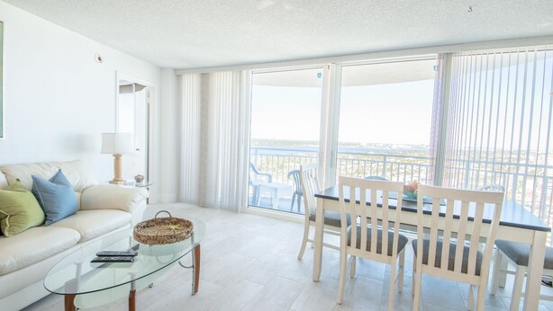 A modern living room with a glass coffee table, a sofa with cushions, a dining area with a table and chairs, and large windows with a view of the sea. - SEA FOREVER 2/2 Great Ocean and River Views Pool & HotTub 20NW (Daytona Beach)