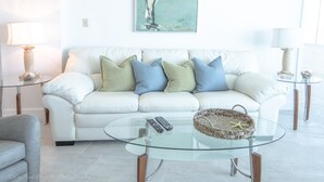 A white sofa with blue and green cushions, a glass coffee table with a starfish, and a painting of a bird on the wall. - SEA FOREVER 2/2 Great Ocean and River Views Pool & HotTub 20NW (Daytona Beach)