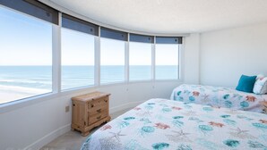 A bedroom with a large window offering a view of the ocean. There are two beds with floral bedding and a wooden nightstand. - SEA FOREVER 2/2 Great Ocean and River Views Pool & HotTub 20NW (Daytona Beach)