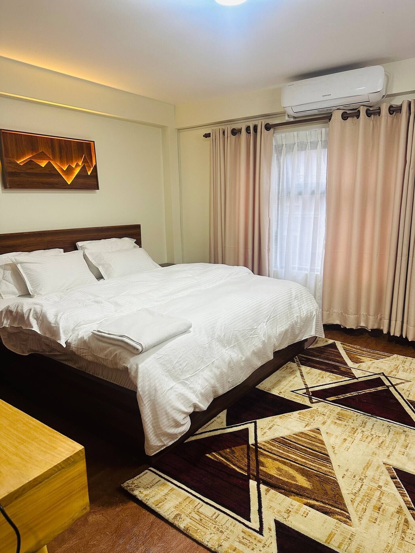 A hotel room with a bed, a wooden headboard, a wall-mounted air conditioner, and a patterned rug.
