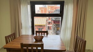 Un comedor con una mesa y sillas de madera, una cesta tejida y vistas de edificios a través de una ventana con cortinas.
