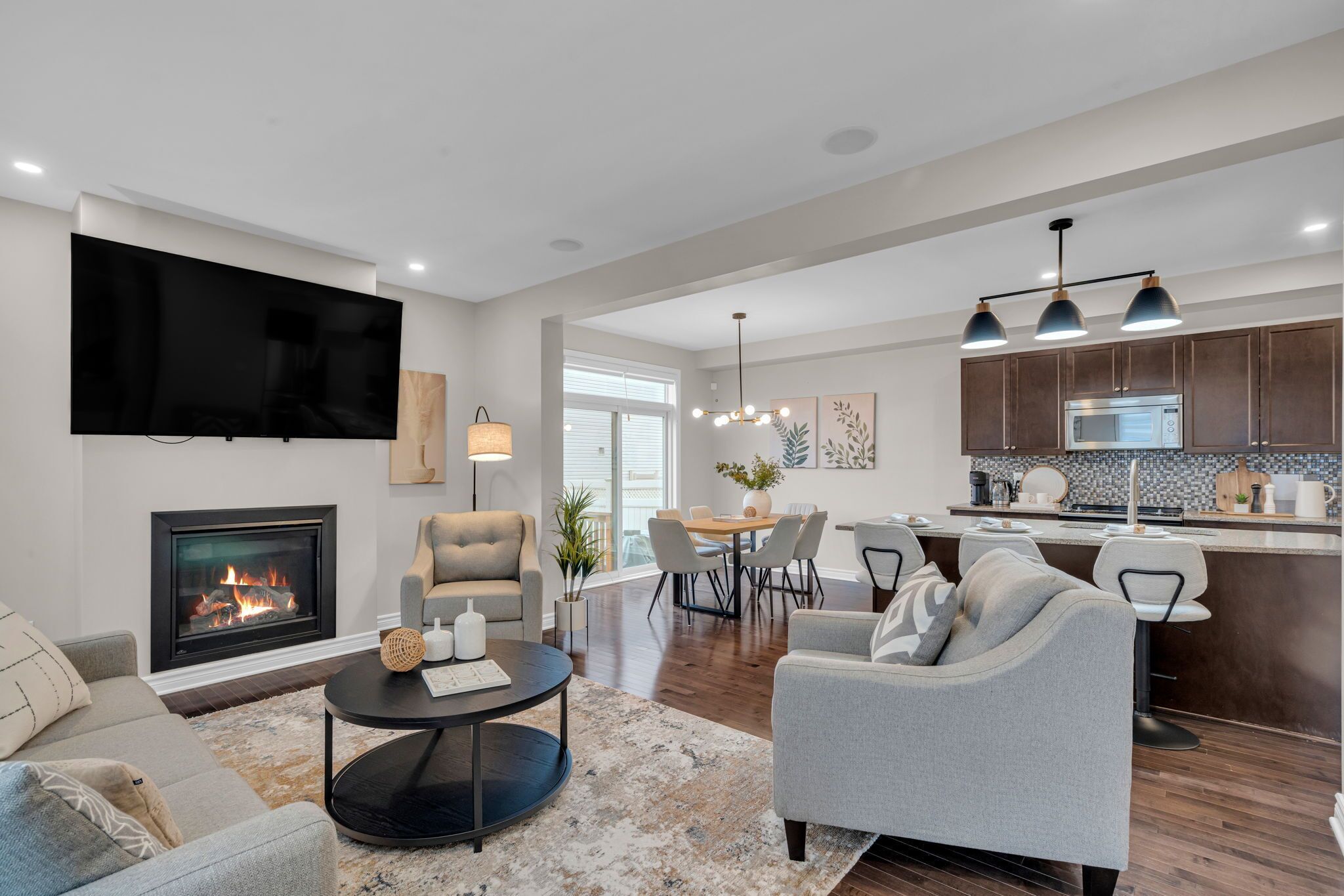A modern living room with a fireplace, a flat-screen TV, and a dining area with a kitchen in the background.