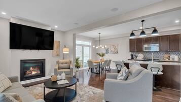 A modern living room with a fireplace, a flat-screen TV, and a dining area with a kitchen in the background.