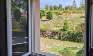 A view through a window of a landscaped outdoor area with a stone wall, various shrubs, and trees. - Gîte du Puech 2-3 personnes (RIEUTORT DE RANDON)