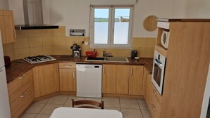 A compact kitchen with wooden cabinets, a white refrigerator, a gas stove, a dishwasher, and a window with a view. - Gîte du Puech 2-3 personnes (RIEUTORT DE RANDON)