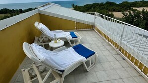 Eine Dachterrasse mit zwei Liegestühlen, einem kleinen runden Tisch mit einer Flasche und Blick auf das Meer und den Strand.