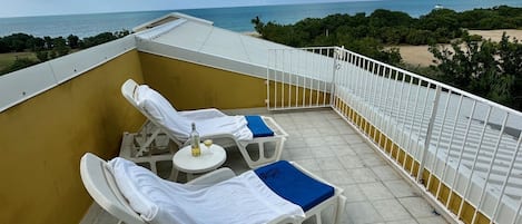 A rooftop terrace with two sun loungers, a small round table with a bottle, and a view of the ocean and beach.