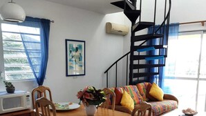 Ein Essbereich mit einem Holztisch und Stühlen, einer Couch mit bunten Kissen, einer Treppe und einem Fenster mit blauen Vorhängen.