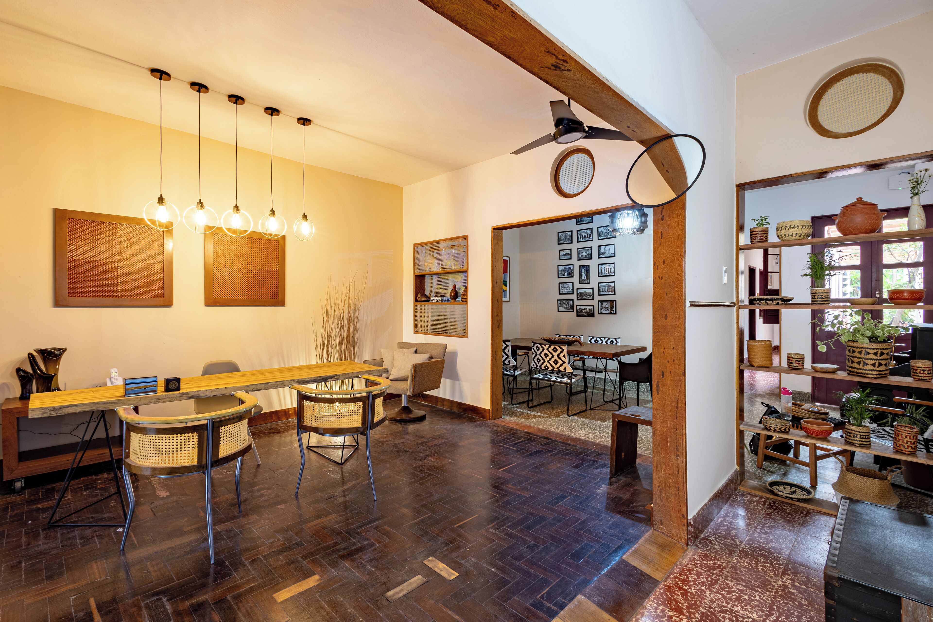 Um quarto espaçoso com piso de madeira, uma mesa comprida com cadeiras, um ventilador de teto e uma parede com quadros.