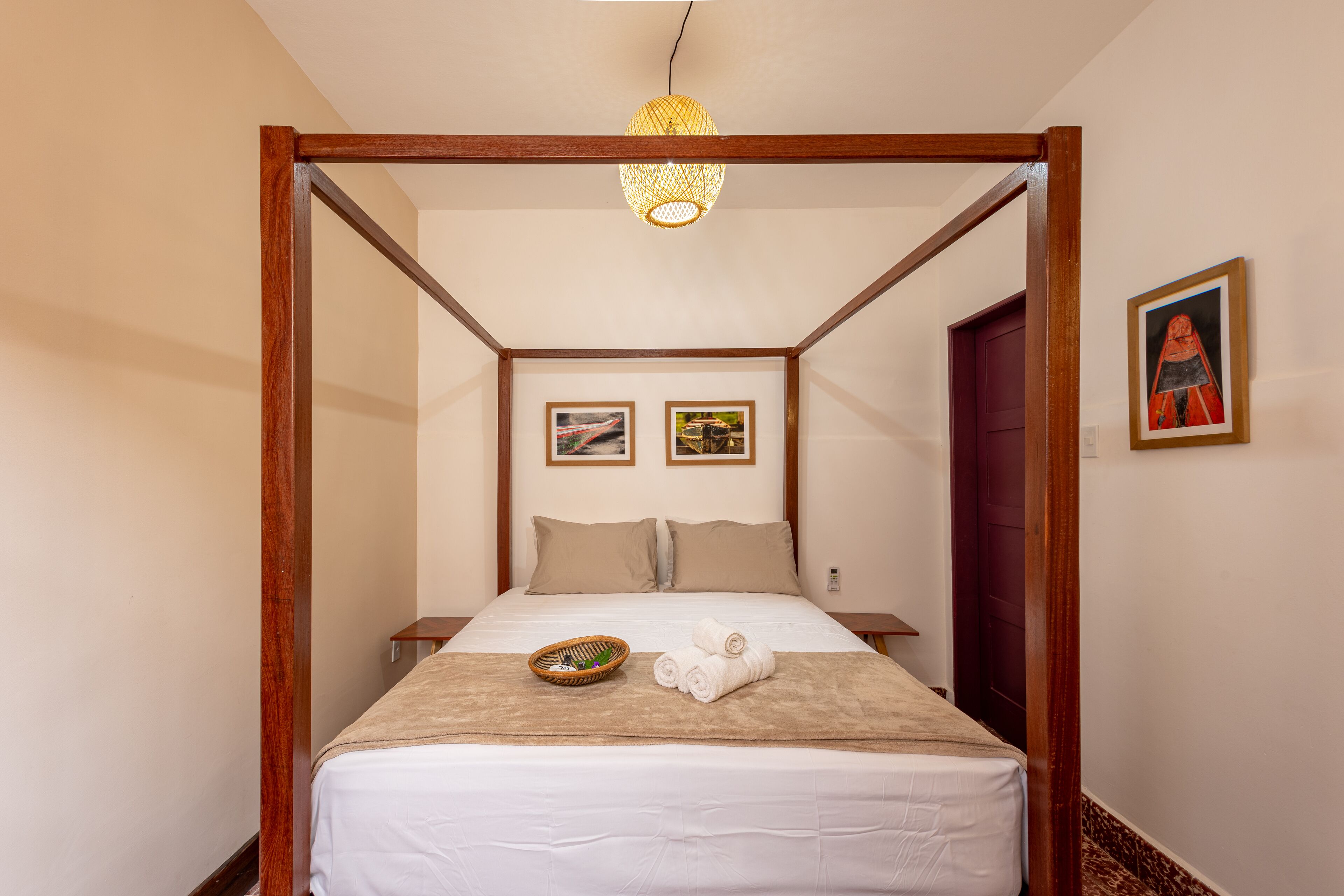Una cama con dosel de madera, una colcha blanca, dos almohadas, una mesita de noche con una lámpara y dos cuadros enmarcados en la pared.