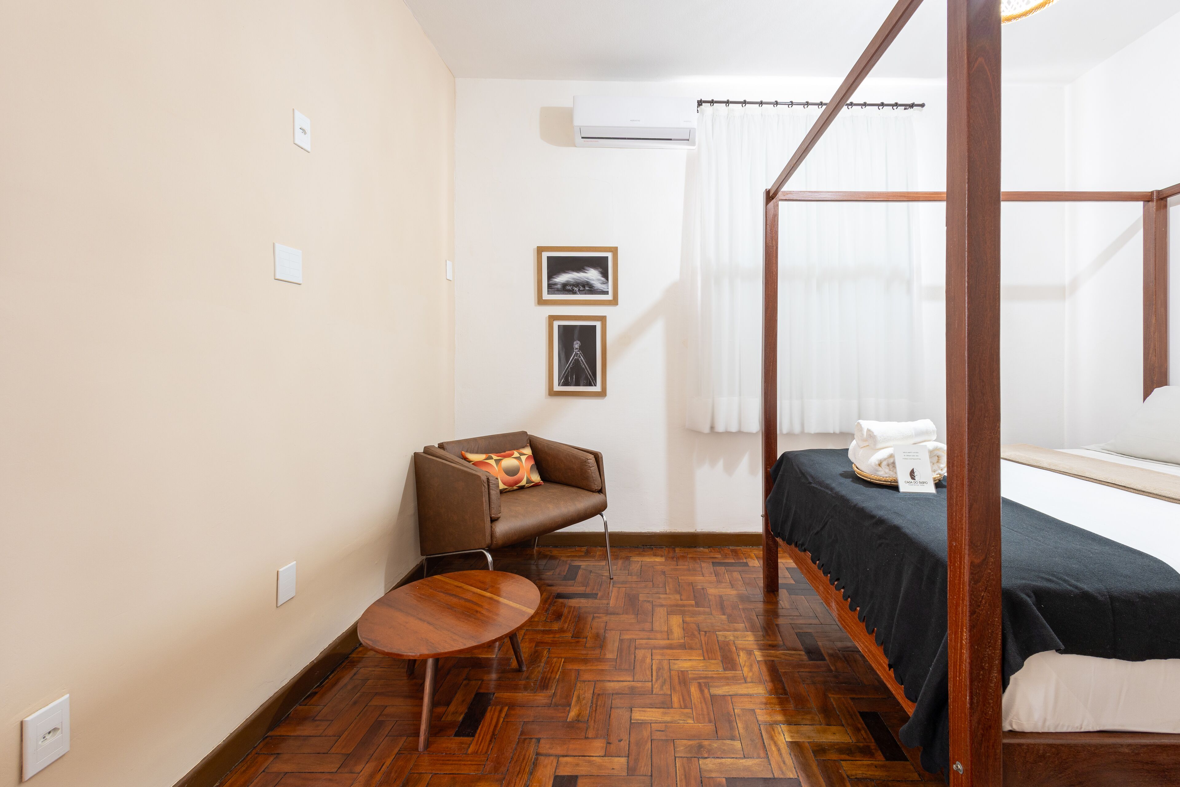 A room with a four-poster bed, a small round table, a brown leather chair, and two framed pictures on the wall.