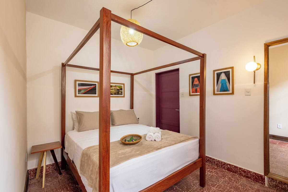 A four-poster bed with a wooden frame, a white bedspread, and a beige throw blanket. There is a small wooden nightstand to the left of the bed.