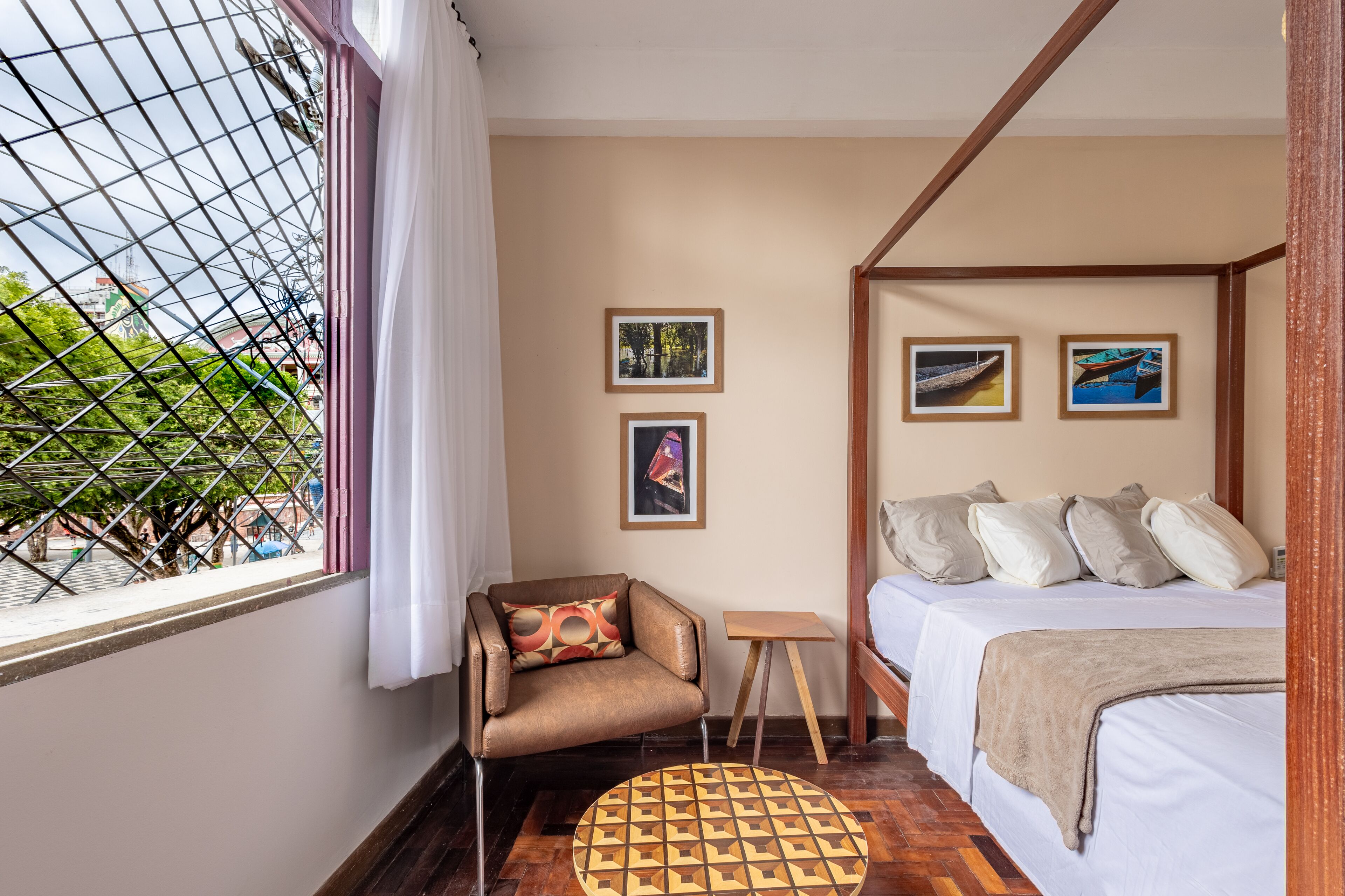 A bedroom with a four-poster bed, a chair with a patterned cushion, a small wooden table, and two framed pictures on the wall.