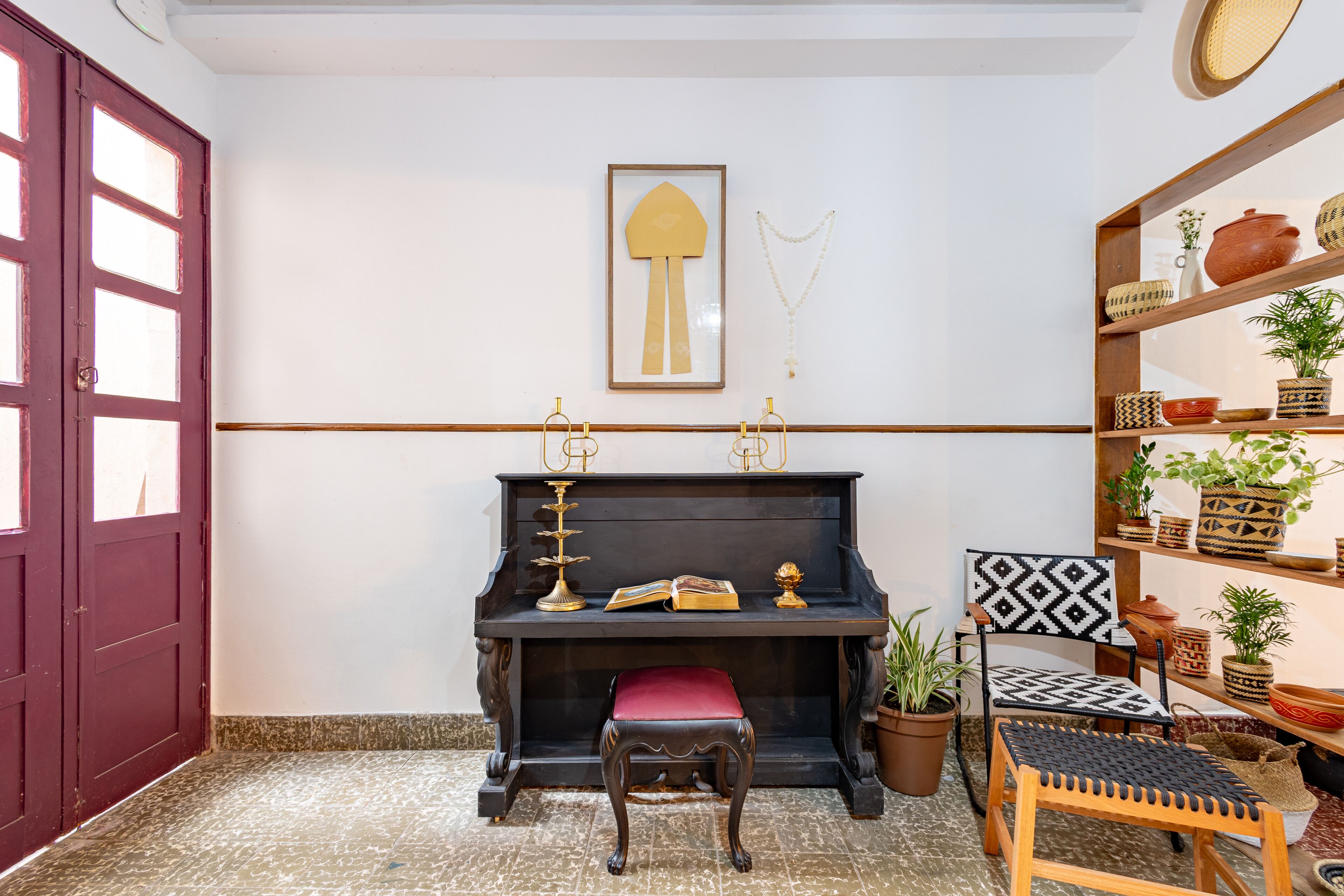 Una habitación con un piano negro, un banco rojo, una silla con respaldo estampado y estantes con plantas en macetas y objetos decorativos.