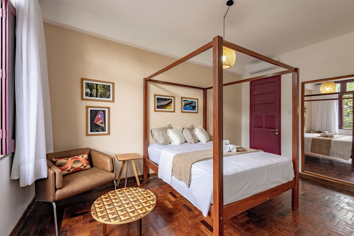 A four-poster bed with a wooden frame, a small round table, a sofa with patterned cushions, and a mirror reflecting the room.