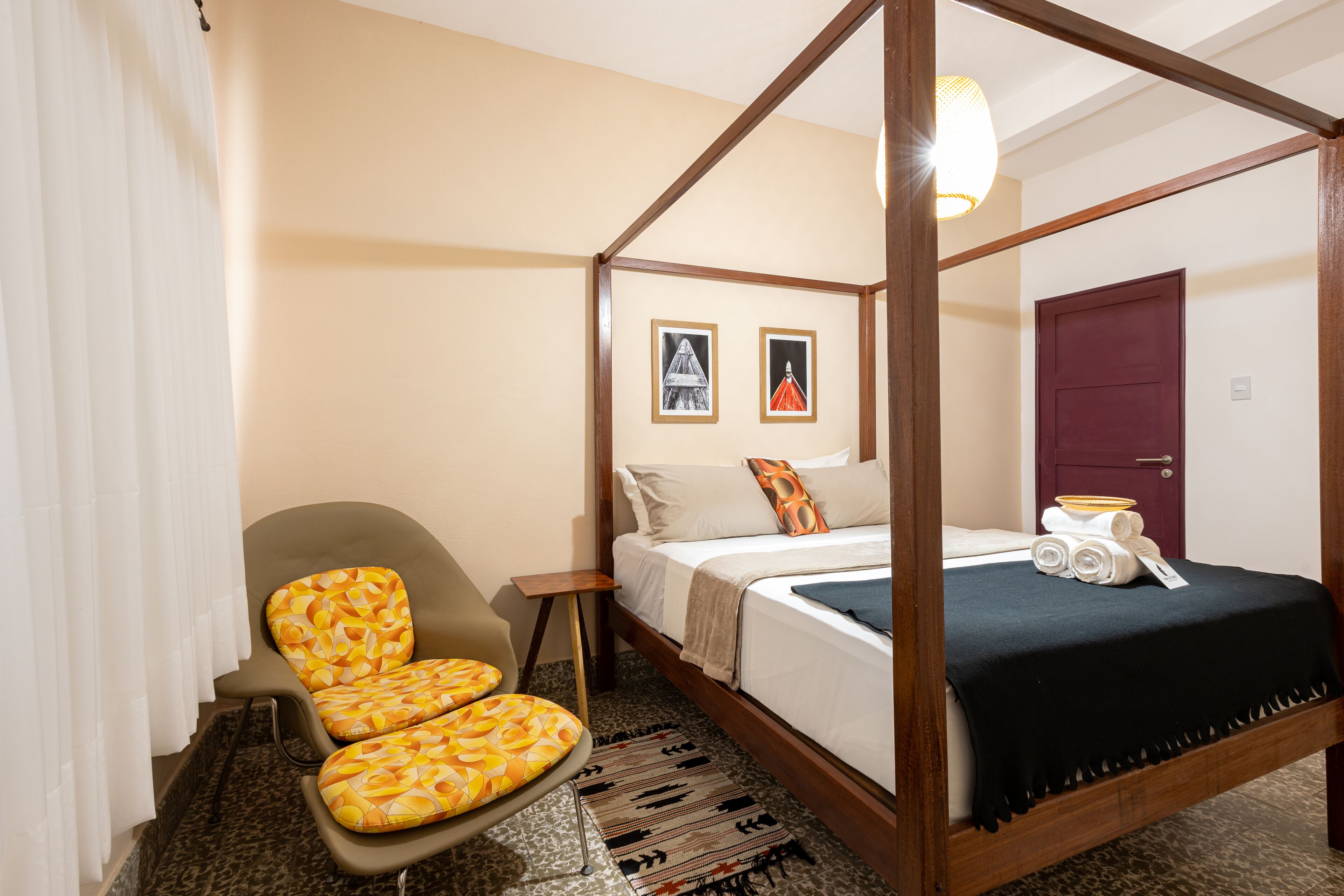 A four-poster bed with a dark bedspread, a chair with a yellow patterned cushion, and a small table with a bowl of towels.