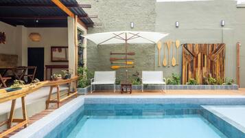 Zona de piscina con dos sillones blancos, una mesa de madera y una sombrilla.