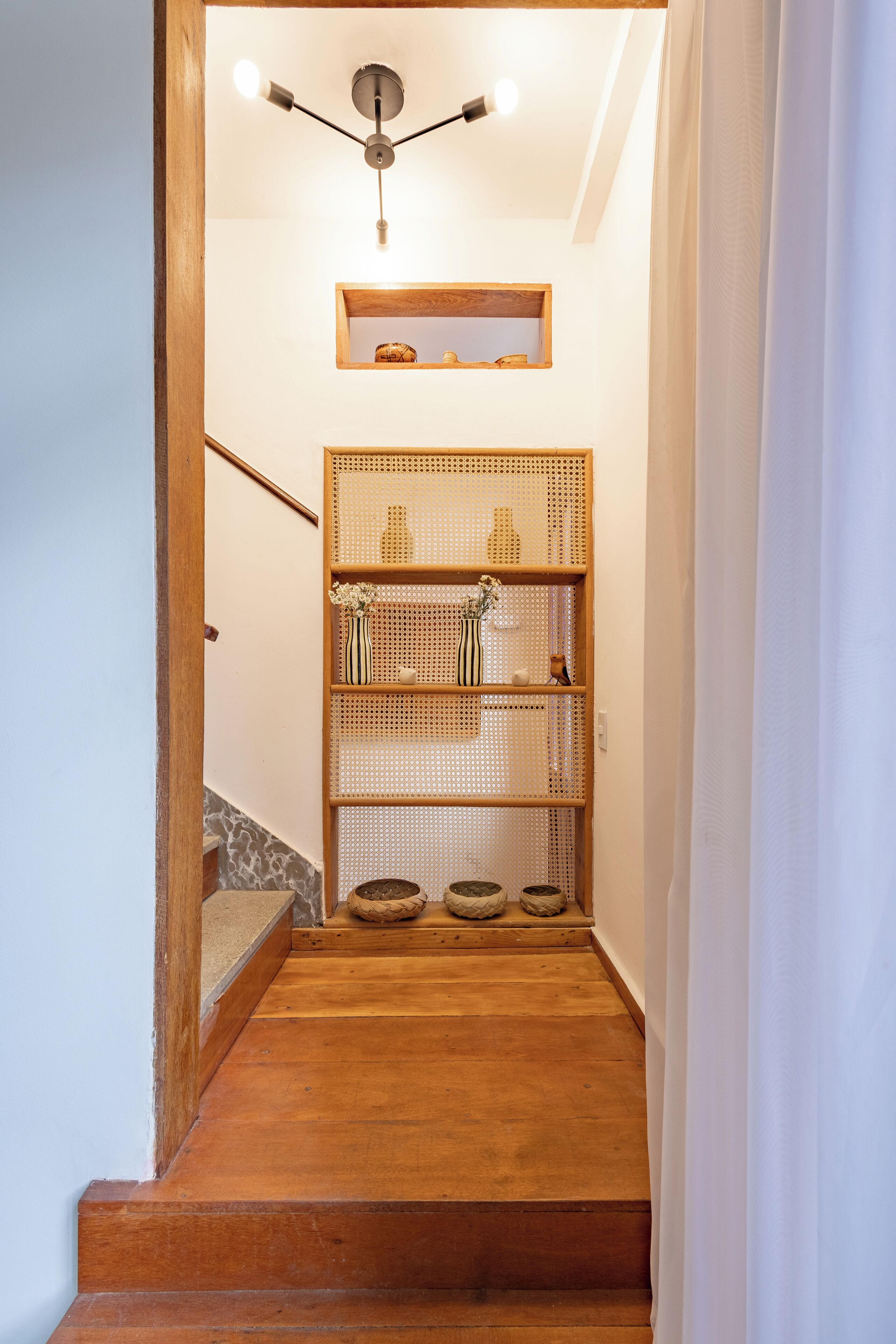 A wooden staircase leading to a landing with a ceiling fan, a woven shelf with decorative items, and a small wooden shelf above.