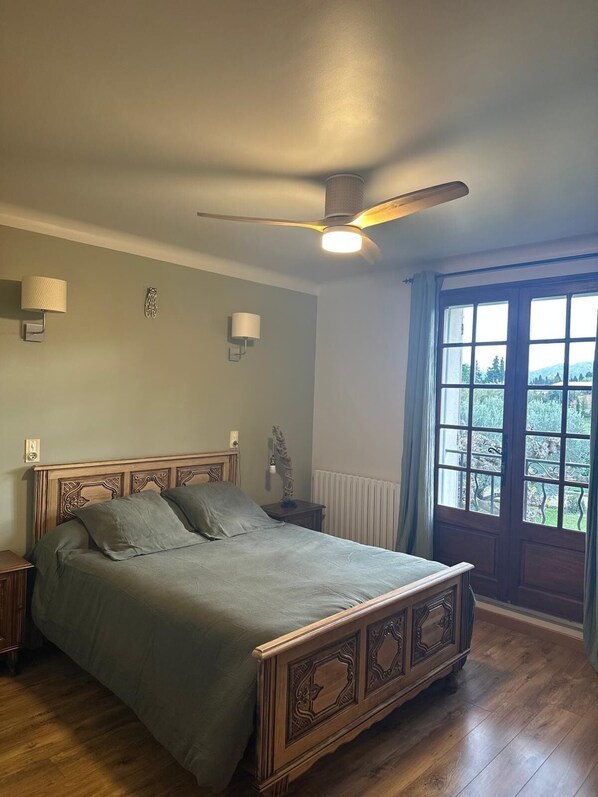 A bedroom with a wooden bed, a ceiling fan, a nightstand, and a window with a view of greenery.