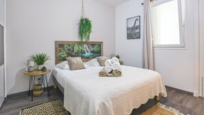 Un dormitorio con una cama, una mesita de noche con una planta, un cuadro enmarcado en la pared y una ventana con cortinas.