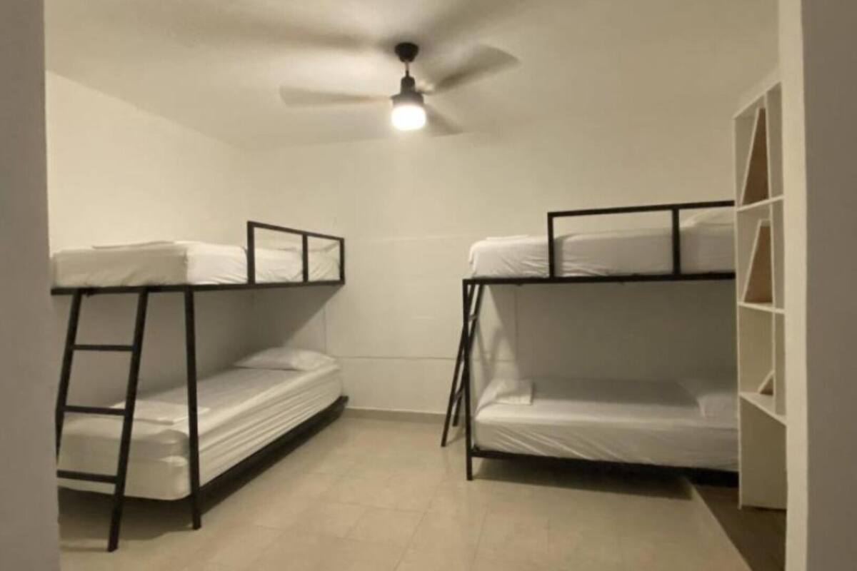 Quarto em dormitório com beliches, ventoinha no teto e piso de azulejos brancos.