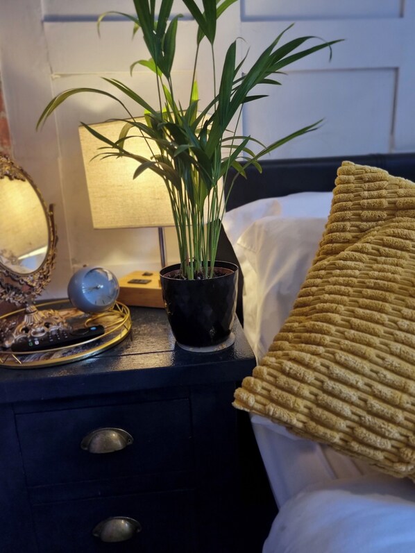 A neatly made bed with a gold throw pillow, a bedside table with a black pot containing a plant, a lamp, and a clock. - Charming 1-bed Studio, Parking, Kitchen,wifi (Hampton)