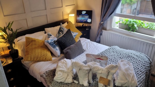 A neatly made bed with a variety of pillows and a knitted blanket, a bedside table with a lamp, a television, and a window with a view of greenery.