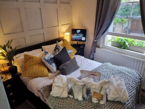 A neatly made bed with a variety of pillows and a knitted blanket, a bedside table with a lamp, a television, and a window with a view of greenery. - Charming 1-bed Studio, Parking, Kitchen,wifi (Hampton)