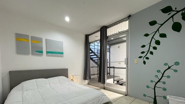 A bedroom with a bed, a window, a door, and a wall with a tree mural. - SJO Oasis Hostel (San José)