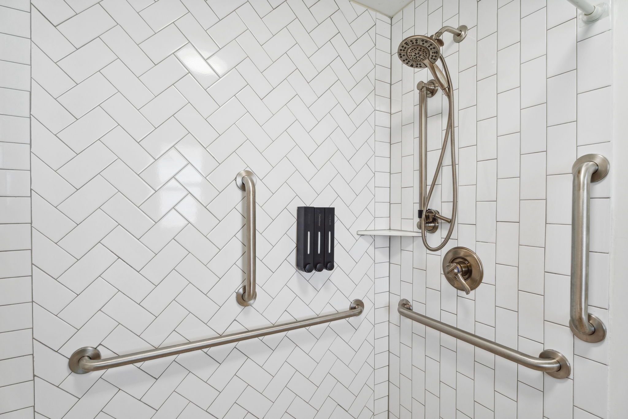 A bathroom with white herringbone tile walls, a shower with a handheld showerhead, and multiple grab bars.