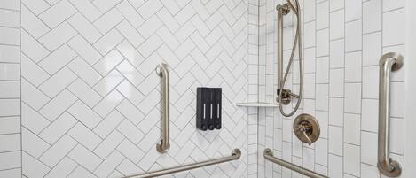 A bathroom with white herringbone tile walls, a shower with a handheld showerhead, and multiple grab bars.