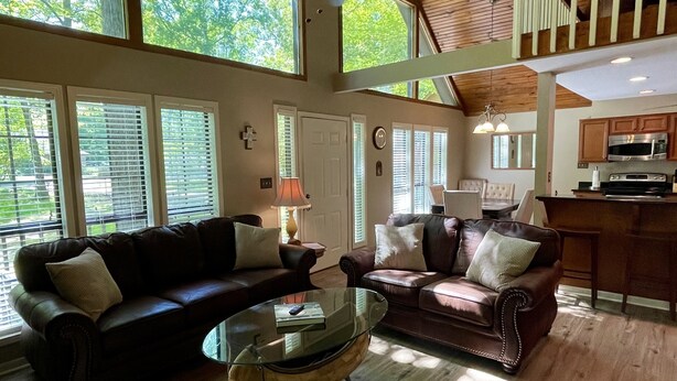 A spacious living room with two leather sofas, a glass coffee table, and a ceiling fan. Large windows allow natural light to fill the space.