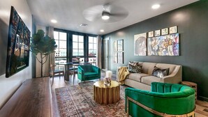 Ein modernes Wohnzimmer mit einem grünen Sofa, einem beigen Sofa und einem runden Couchtisch. Große Fenster, ein Deckenventilator und Kunstwerke schmücken die Wände.