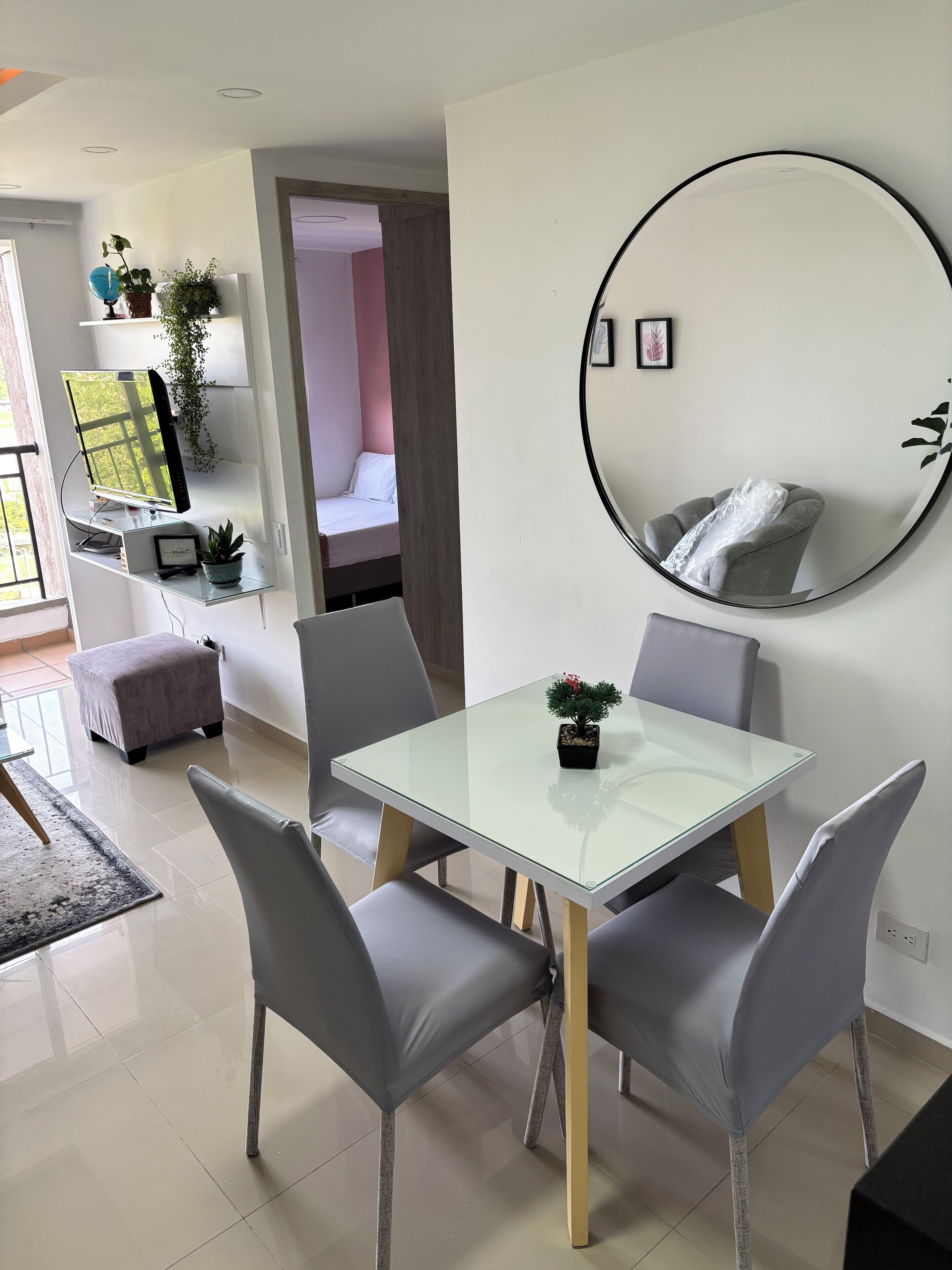 A modern living room with a glass dining table, grey upholstered chairs, a round mirror, and a TV on a stand.