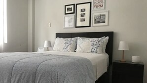 A neatly made bed with a grey quilt, two white pillows with a leaf pattern, and a dark headboard. There are two bedside tables with lamps and framed pictures on the wall above. - InkaHuset Airport (Callao)