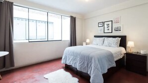 A bedroom with a bed, a nightstand, a lamp, a window with curtains, and a wall with framed pictures. - InkaHuset Airport (Callao)