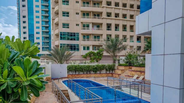 Piscina no terraço com grade de metal, rodeada por vegetação e um edifício alto ao fundo.