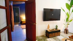 Une pièce avec une porte en bois, un téléviseur fixé au mur, un petit meuble en bois orné d’objets décoratifs et une plante en pot.