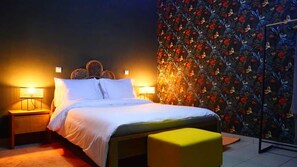 A bed with white bedding, two bedside lamps, a wooden nightstand, a yellow ottoman, and a floral wallpaper.