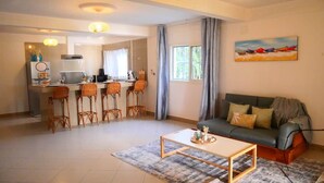 A living room with a sofa, a coffee table, and a kitchen area with a dining table and chairs. - Studio Océan - Vue sur Mer/Pied dans l'eau (Libreville)
